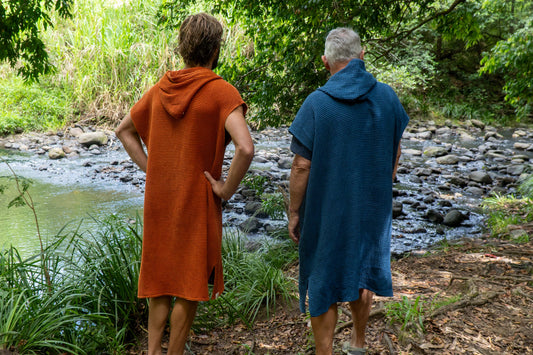 Two people in hooded towels standing by a stream in a forested area